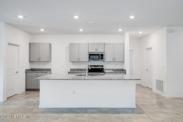 kitchen with gray cabinetry, light stone countertops, stainless steel appliances, and a center island with sink