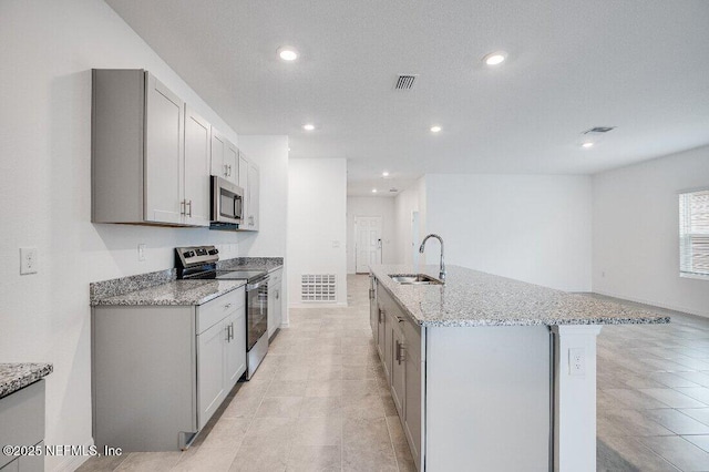 kitchen featuring gray cabinets, appliances with stainless steel finishes, sink, light stone counters, and a center island with sink