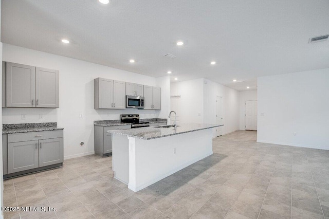 kitchen with gray cabinets, sink, a kitchen island with sink, light stone counters, and stainless steel appliances