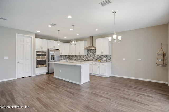 kitchen with light wood-type flooring, wall chimney exhaust hood, stainless steel appliances, a kitchen island with sink, and hanging light fixtures