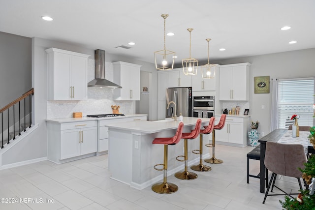 kitchen with stainless steel appliances, a kitchen island with sink, wall chimney range hood, decorative light fixtures, and white cabinets