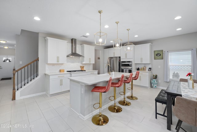 kitchen featuring a center island with sink, wall chimney exhaust hood, decorative light fixtures, white cabinetry, and stainless steel appliances