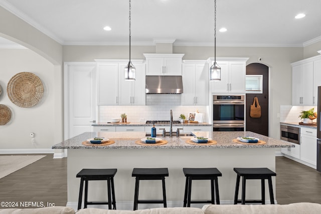 kitchen with a kitchen island with sink, dark wood-type flooring, hanging light fixtures, ornamental molding, and light stone countertops