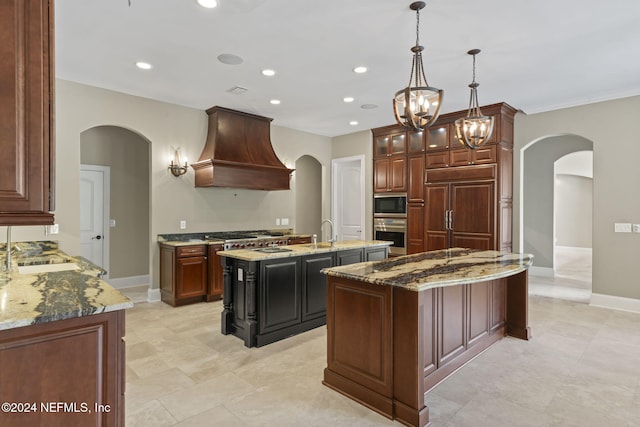 kitchen featuring light stone countertops, custom range hood, built in appliances, a center island with sink, and a chandelier