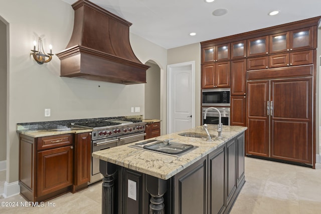 kitchen with premium range hood, light stone counters, sink, built in appliances, and an island with sink