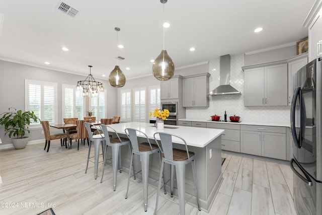 kitchen featuring wall chimney range hood, black fridge, an island with sink, decorative light fixtures, and decorative backsplash