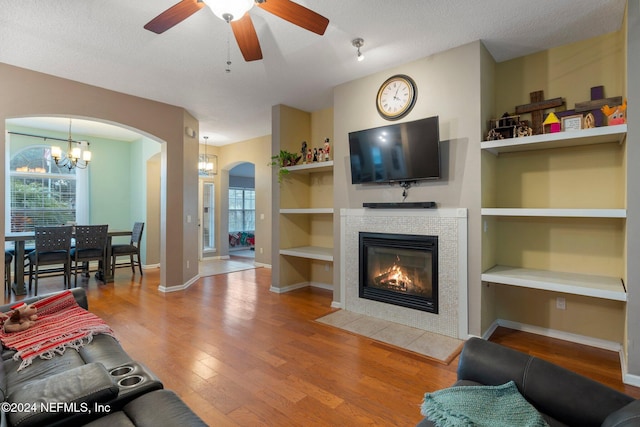 living room featuring built in shelves, a textured ceiling, ceiling fan with notable chandelier, a fireplace, and hardwood / wood-style floors