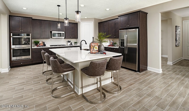 kitchen with pendant lighting, an island with sink, dark brown cabinets, light hardwood / wood-style floors, and stainless steel appliances