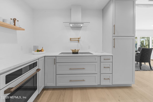 kitchen with gray cabinetry, oven, black electric stovetop, wall chimney range hood, and light wood-type flooring