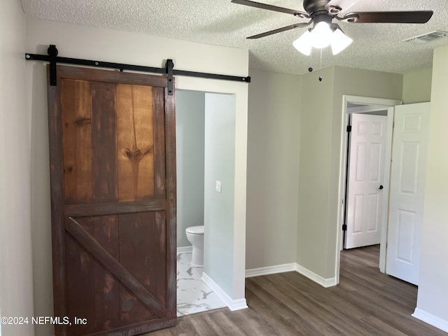 unfurnished bedroom with connected bathroom, ceiling fan, a barn door, dark hardwood / wood-style floors, and a textured ceiling