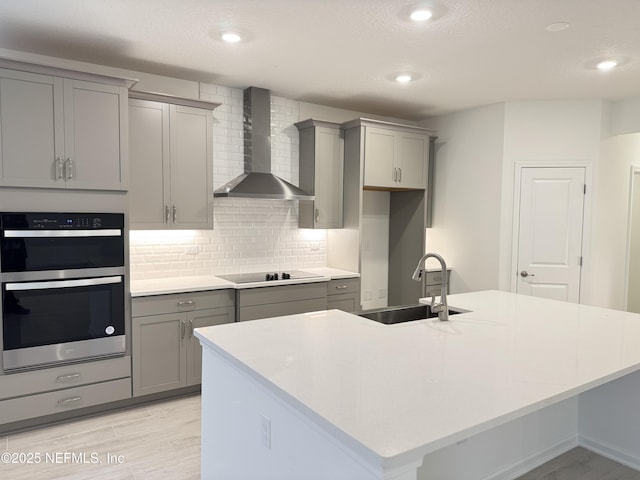 kitchen with wall chimney range hood, gray cabinets, stainless steel double oven, and a sink