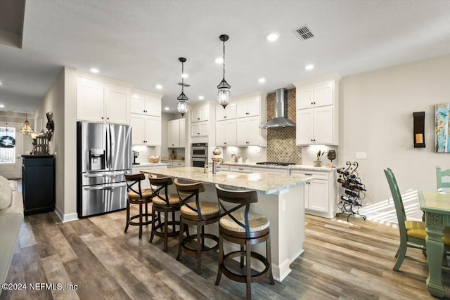 kitchen featuring pendant lighting, a center island with sink, wall chimney range hood, appliances with stainless steel finishes, and white cabinetry