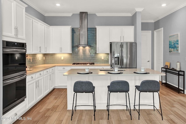 kitchen with appliances with stainless steel finishes, wall chimney exhaust hood, a kitchen island with sink, white cabinets, and light hardwood / wood-style floors