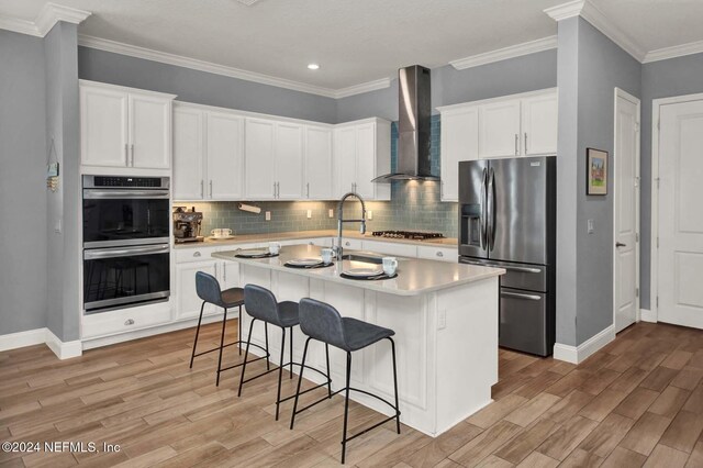 kitchen featuring wall chimney exhaust hood, light wood-type flooring, an island with sink, white cabinetry, and stainless steel appliances