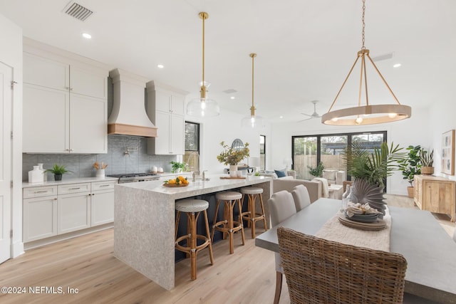 kitchen with pendant lighting, a kitchen island with sink, white cabinets, light hardwood / wood-style flooring, and custom range hood