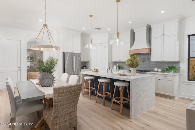 kitchen with stainless steel fridge, decorative light fixtures, a center island with sink, custom range hood, and light wood-type flooring