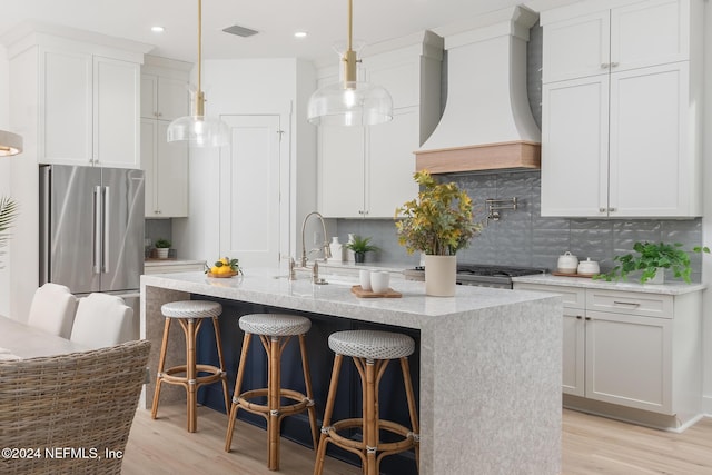 kitchen with high end refrigerator, custom exhaust hood, a center island with sink, light hardwood / wood-style flooring, and white cabinetry