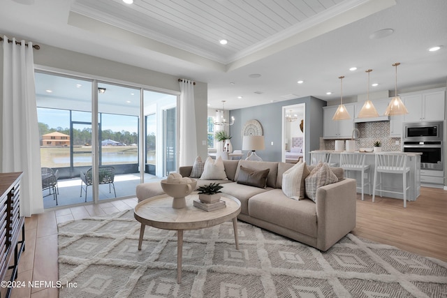 living room with crown molding, a chandelier, a tray ceiling, a water view, and light wood-type flooring