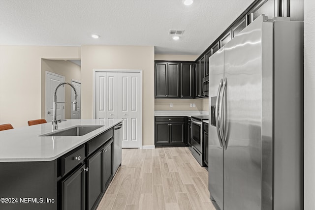 kitchen with appliances with stainless steel finishes, light wood-type flooring, a textured ceiling, sink, and a center island with sink