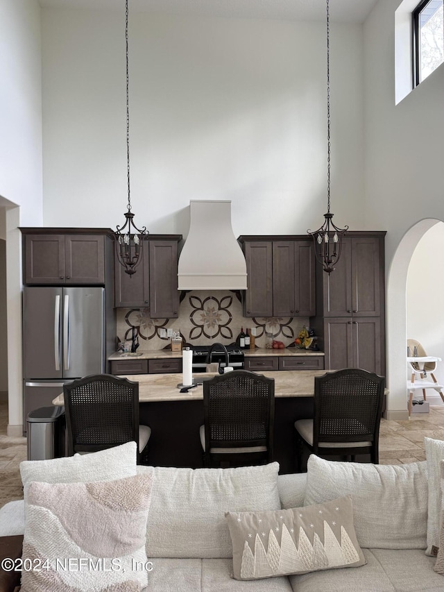 kitchen featuring tasteful backsplash, custom range hood, pendant lighting, a high ceiling, and stainless steel refrigerator