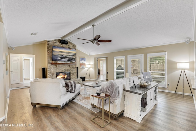 living room with ceiling fan, a brick fireplace, lofted ceiling with beams, light hardwood / wood-style flooring, and a textured ceiling