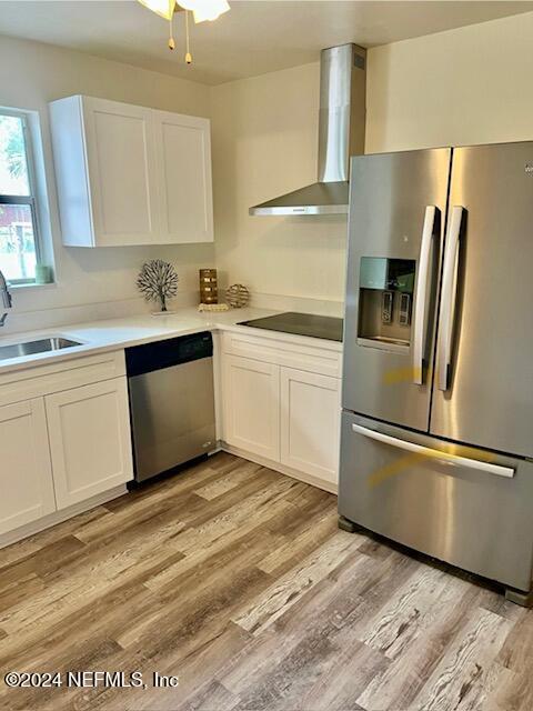 kitchen with white cabinets, wall chimney range hood, sink, appliances with stainless steel finishes, and light hardwood / wood-style floors