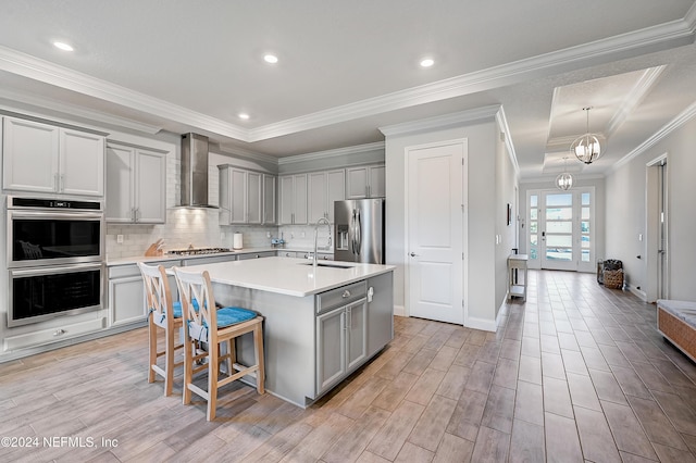 kitchen with a breakfast bar, a kitchen island with sink, wall chimney range hood, crown molding, and appliances with stainless steel finishes