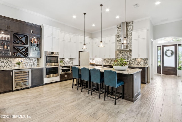 kitchen with stainless steel appliances, beverage cooler, wall chimney range hood, a center island with sink, and hanging light fixtures