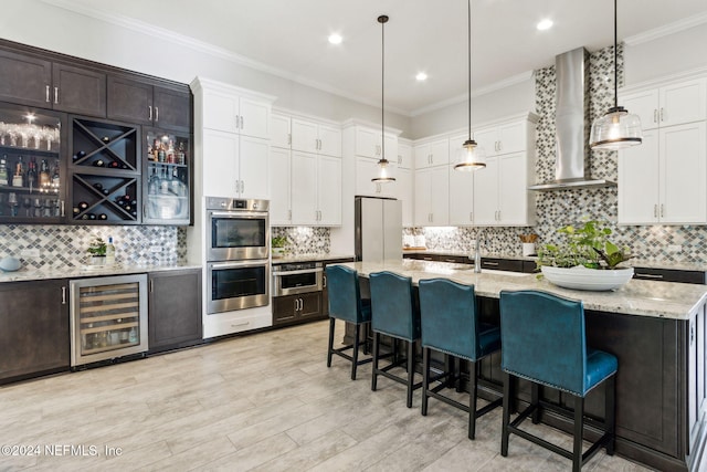 kitchen featuring stainless steel appliances, wall chimney range hood, pendant lighting, wine cooler, and an island with sink