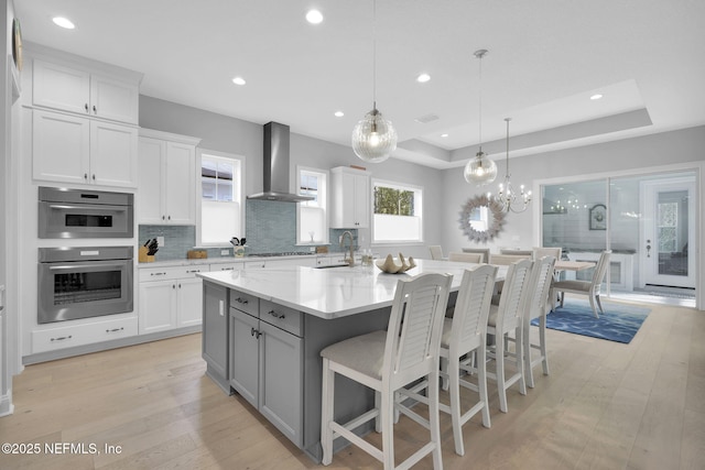 kitchen with white cabinets, a raised ceiling, wall chimney exhaust hood, and a kitchen island with sink