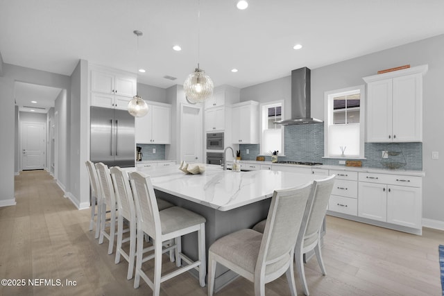 kitchen with appliances with stainless steel finishes, hanging light fixtures, wall chimney range hood, white cabinets, and a large island with sink