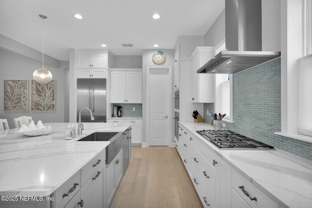 kitchen with pendant lighting, appliances with stainless steel finishes, wall chimney exhaust hood, white cabinetry, and light stone countertops