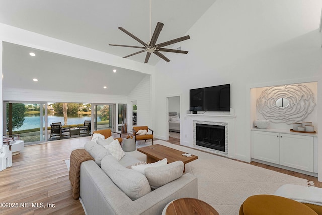 living room featuring high vaulted ceiling, a fireplace, ceiling fan, a water view, and light hardwood / wood-style flooring