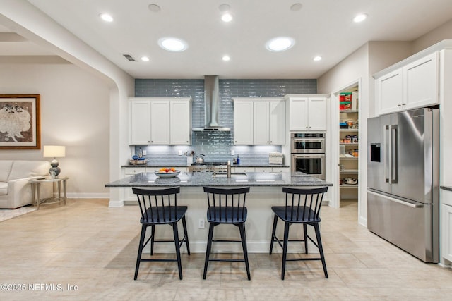 kitchen with white cabinetry, a kitchen island with sink, stainless steel appliances, and wall chimney range hood