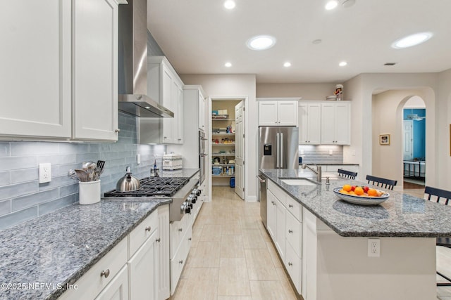 kitchen with wall chimney range hood, an island with sink, a kitchen bar, white cabinetry, and stainless steel appliances