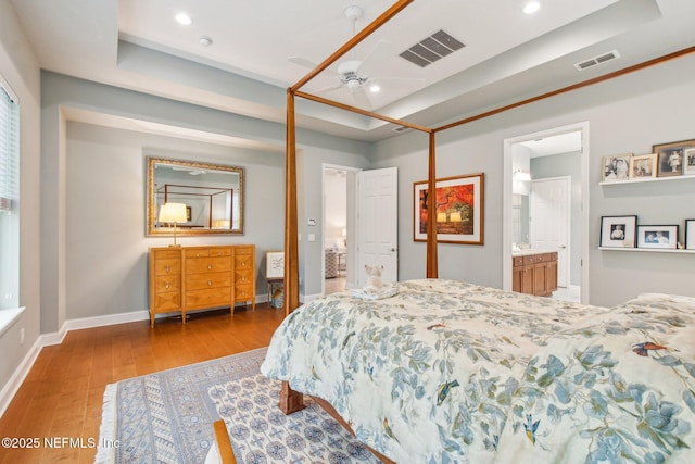 bedroom featuring a raised ceiling, ensuite bath, ceiling fan, and light hardwood / wood-style flooring