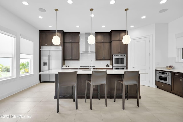 kitchen featuring wall chimney exhaust hood, an island with sink, decorative light fixtures, and appliances with stainless steel finishes