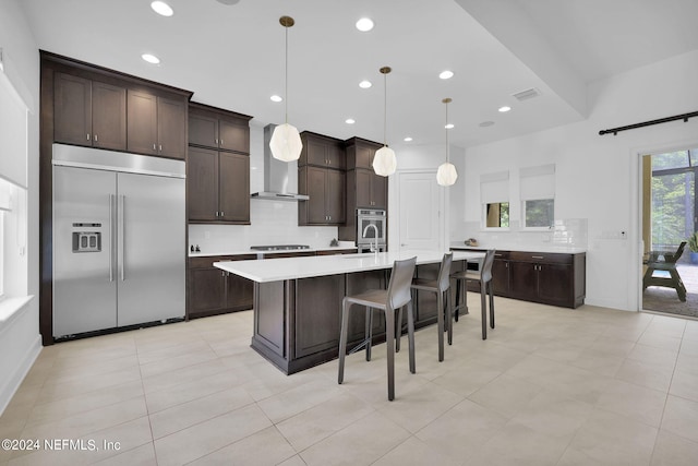 kitchen with dark brown cabinetry, wall chimney exhaust hood, pendant lighting, a kitchen island with sink, and appliances with stainless steel finishes