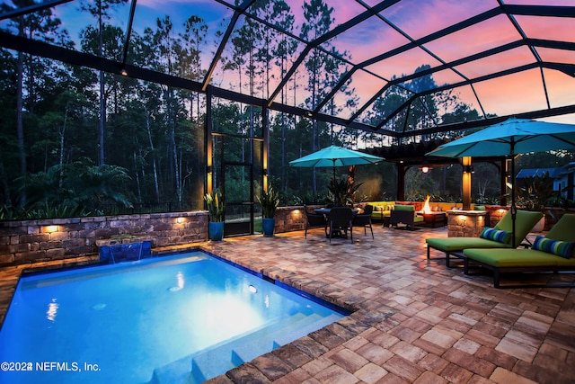 pool at dusk featuring pool water feature, glass enclosure, an outdoor living space with a fire pit, and a patio area