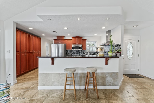 kitchen with kitchen peninsula, a breakfast bar area, appliances with stainless steel finishes, and wall chimney range hood