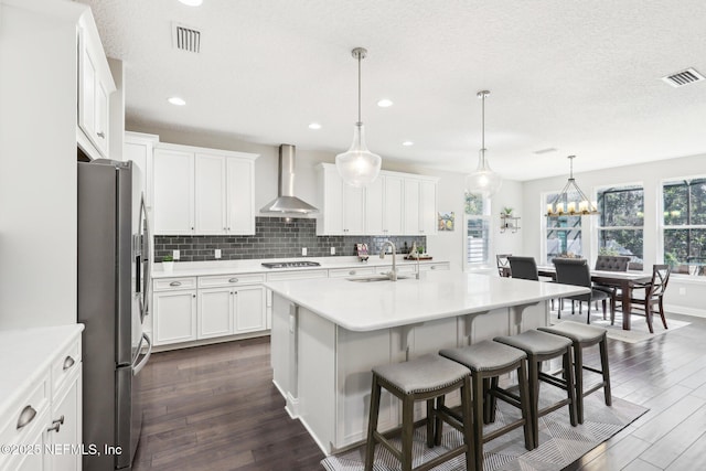 kitchen with hanging light fixtures, wall chimney range hood, sink, stainless steel appliances, and a center island with sink