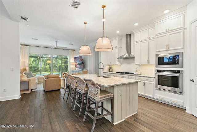 kitchen with white cabinetry, decorative light fixtures, appliances with stainless steel finishes, an island with sink, and wall chimney range hood