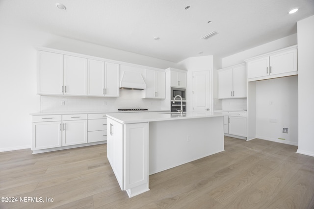 kitchen featuring a kitchen island with sink, premium range hood, sink, light wood-type flooring, and white cabinetry