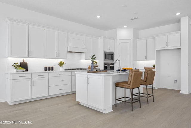 kitchen featuring white cabinets, custom exhaust hood, an island with sink, light hardwood / wood-style floors, and stainless steel microwave