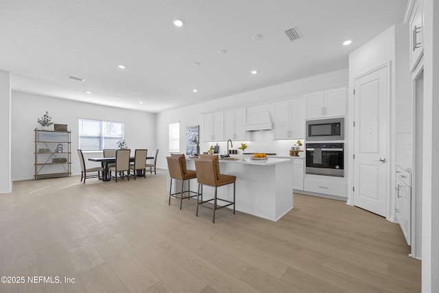 kitchen featuring white cabinets, custom exhaust hood, an island with sink, oven, and built in microwave