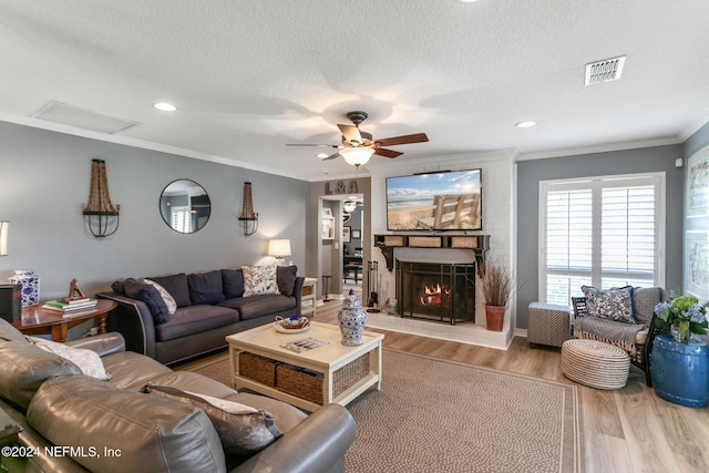 living room with ceiling fan, a brick fireplace, crown molding, a textured ceiling, and light wood-type flooring