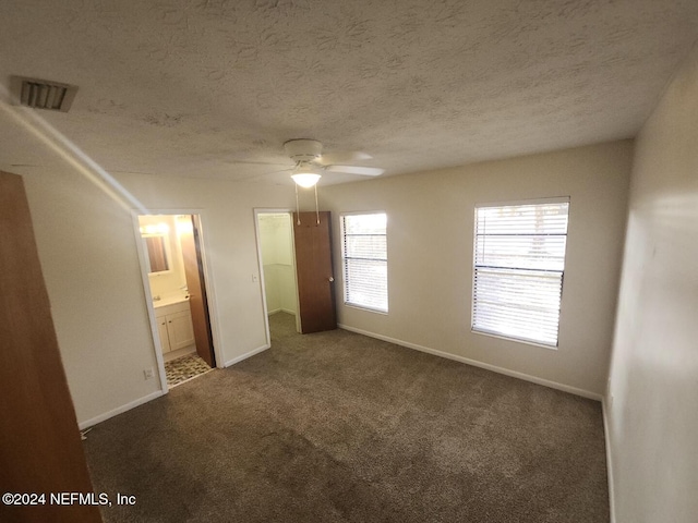 unfurnished bedroom with a textured ceiling, visible vents, baseboards, dark carpet, and ensuite bath