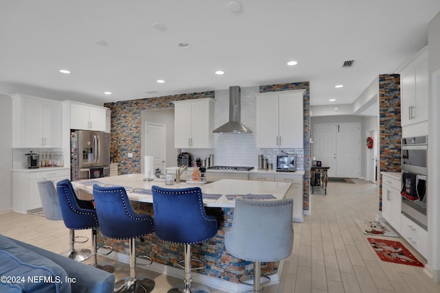 kitchen with a kitchen island with sink, backsplash, white cabinets, wall chimney exhaust hood, and appliances with stainless steel finishes