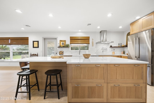 kitchen featuring high end fridge, wall chimney exhaust hood, sink, a kitchen island, and a breakfast bar area