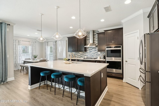 kitchen with pendant lighting, sink, an island with sink, stainless steel appliances, and wall chimney exhaust hood